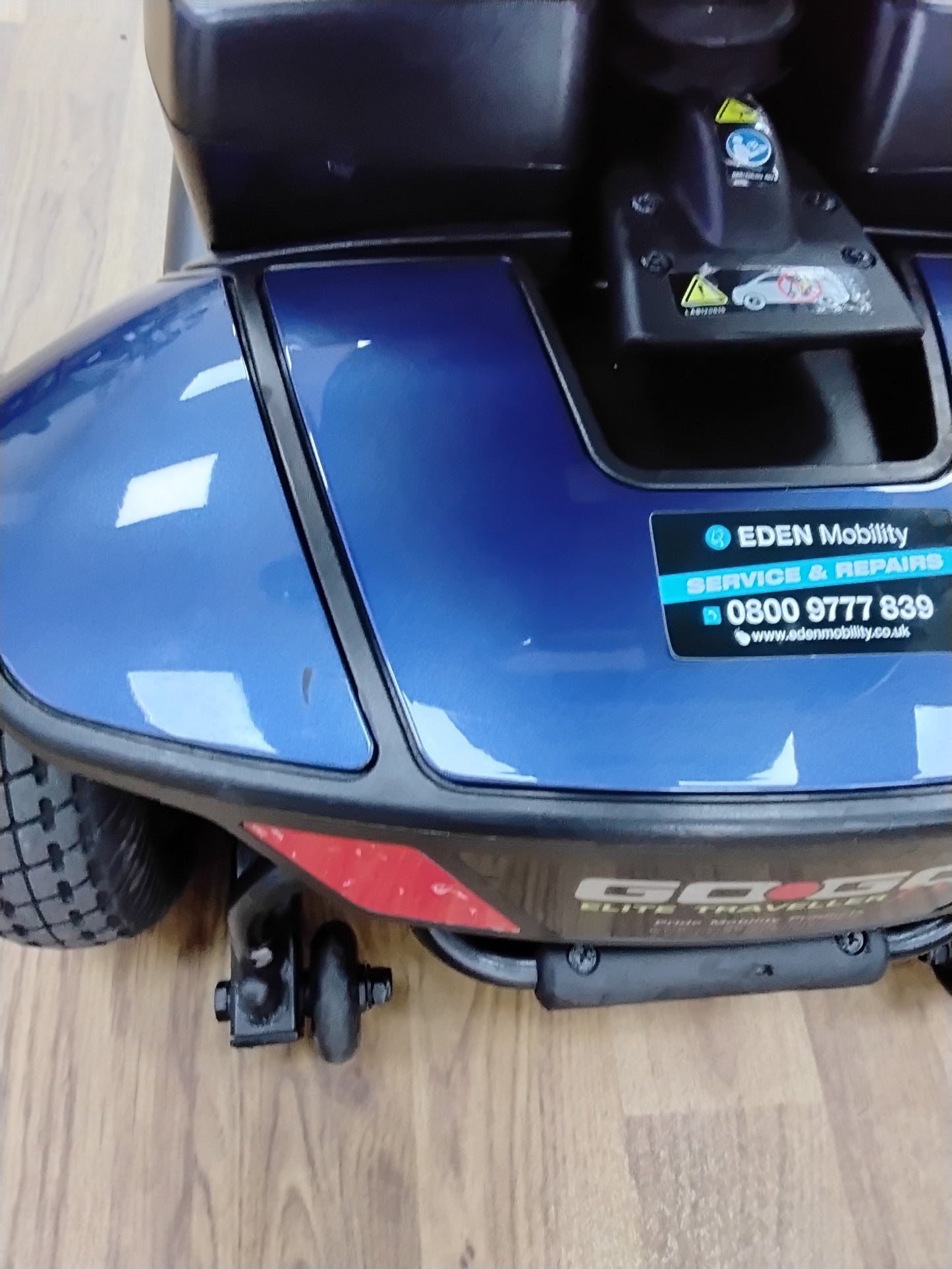 Close-up of the blue Pride Go Go Elite Traveller mobility scooter with Eden Mobility service sticker on wooden floor background
