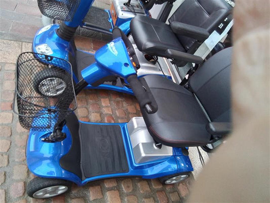 Blue Kymco Mini Comfort mobility scooter with black seat, armrests, front basket, and four wheels on cobblestone ground