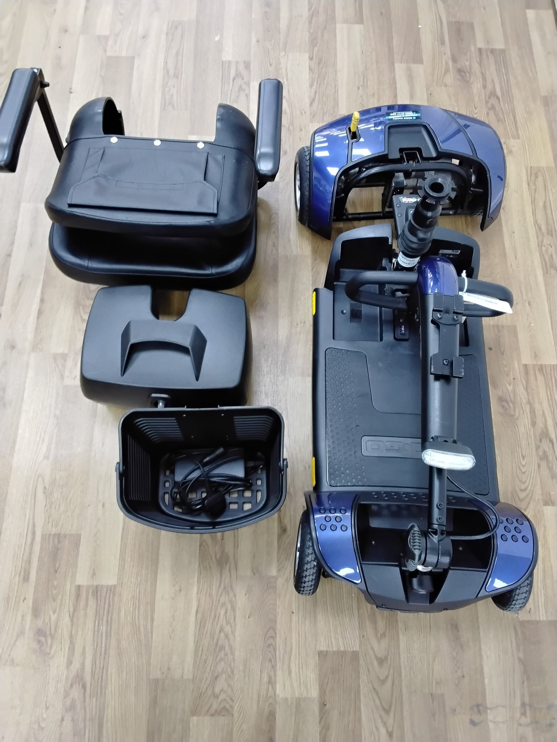 Disassembled Pride Go Go Elite Traveller mobility scooter in blue, showing seat, battery cover, and basket on wooden floor.