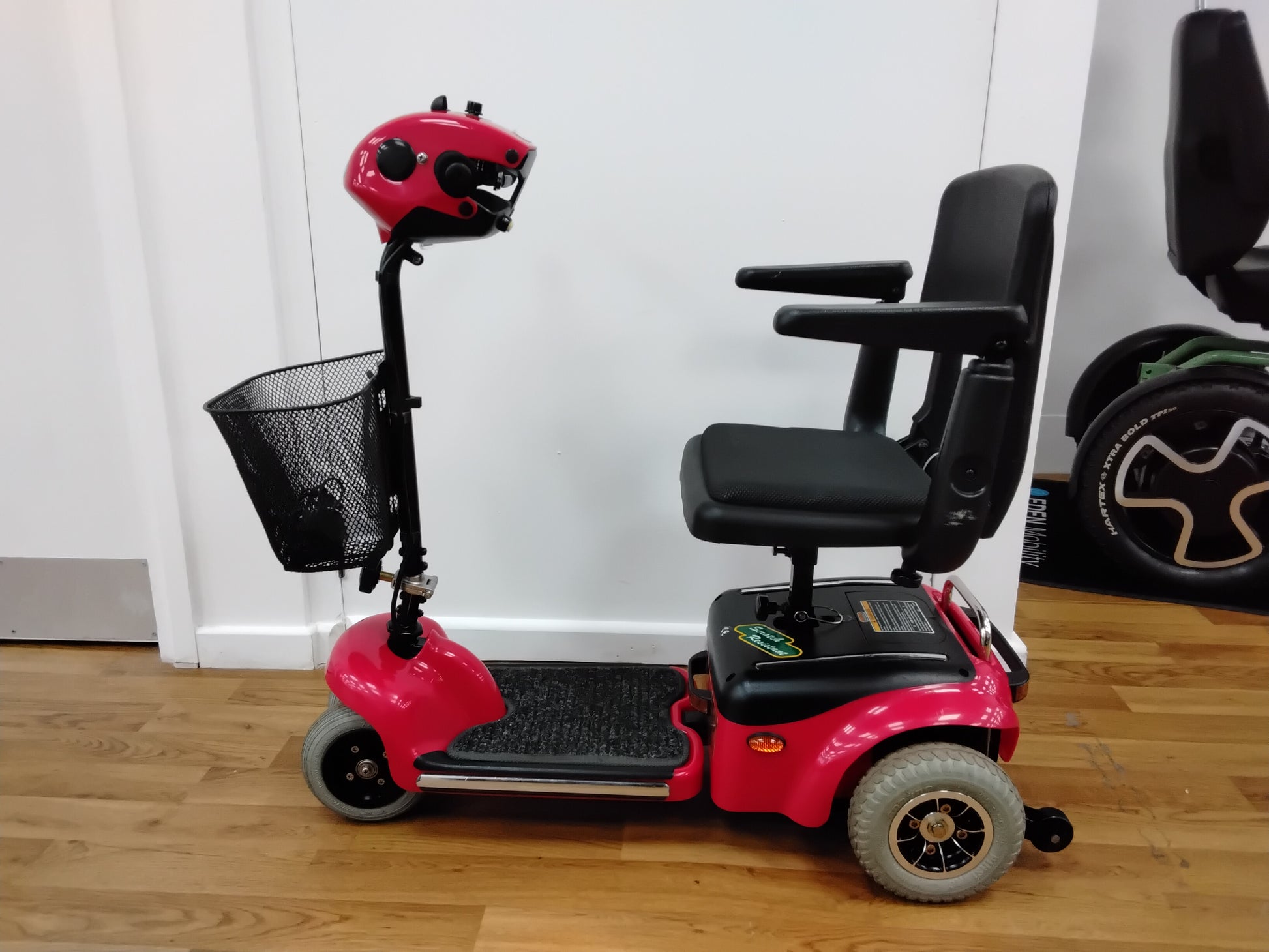 Red Shoprider Whisper mobility scooter with black seat, armrests, and front mesh basket on wooden floor indoors