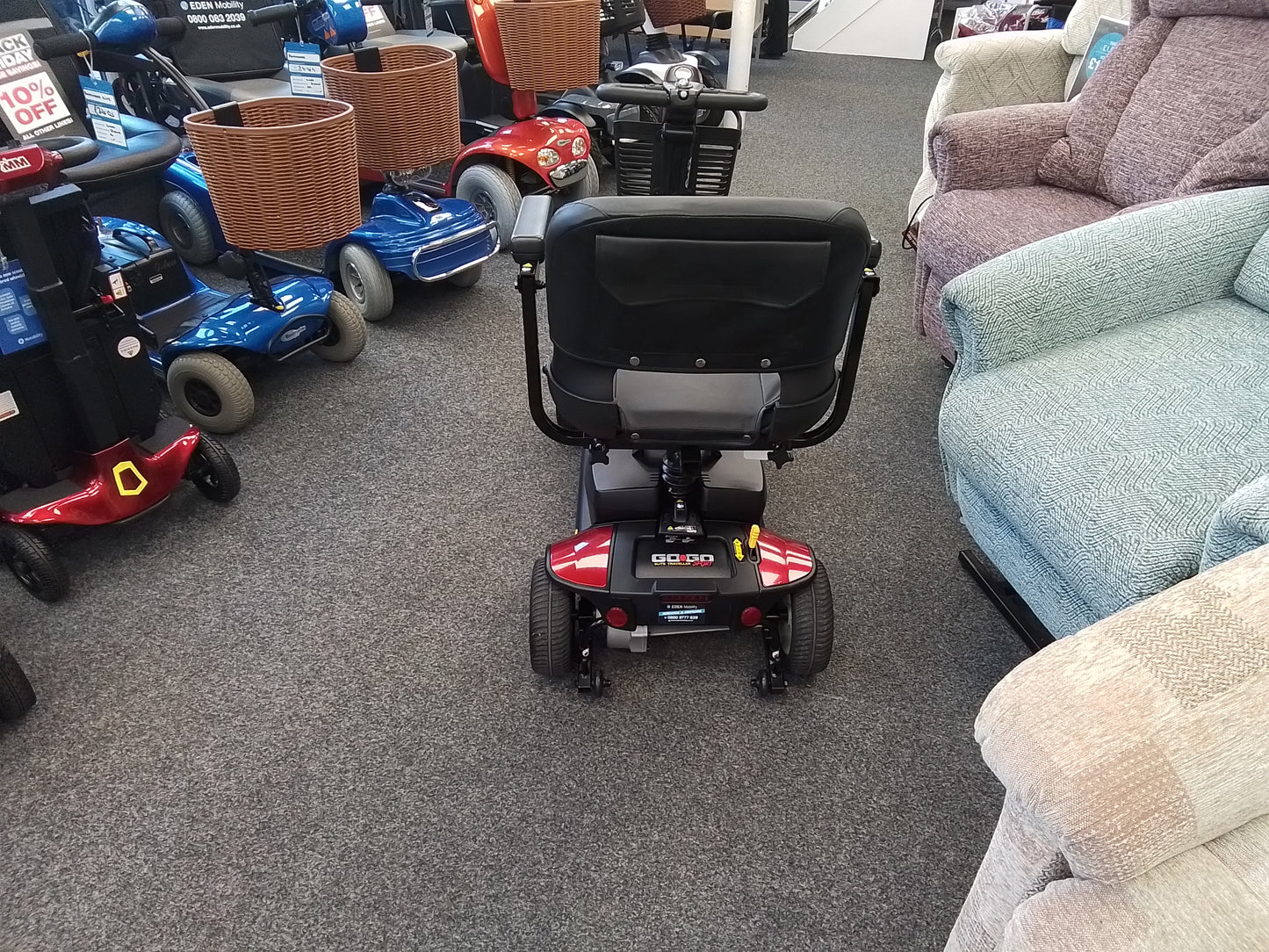Rear view of a red Pride Go Go Elite Traveller mobility scooter displayed indoors on a gray carpet among chairs and scooters