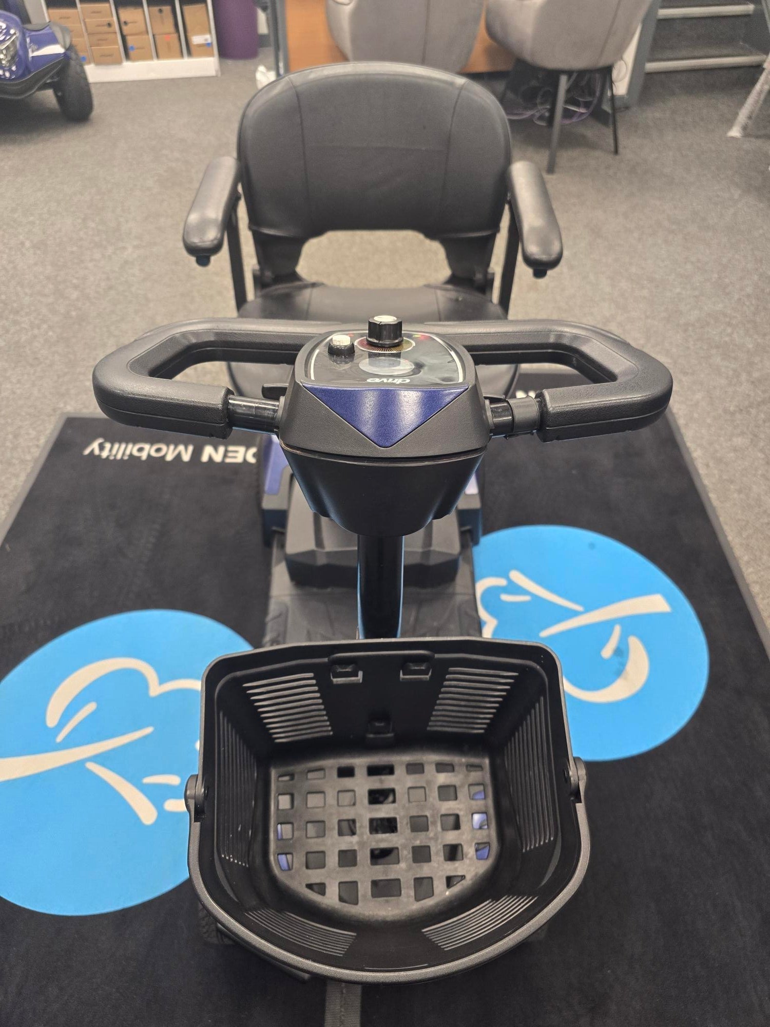 Close-up of a refurbished blue Drive Scout mobility scooter with handlebar controls and front basket on display indoors.