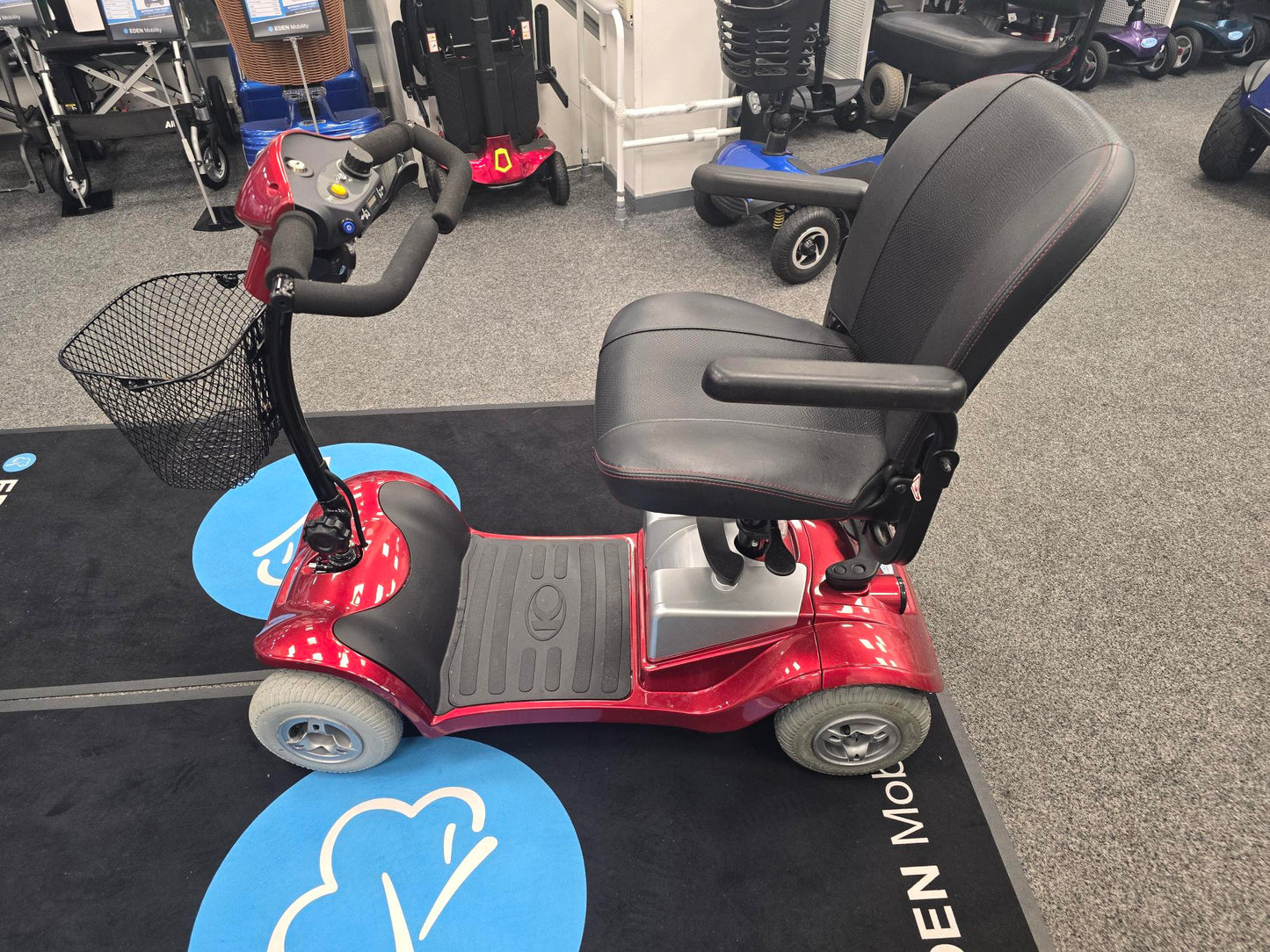 Red Kymco Mini Comfort mobility scooter with black seat and basket on gray carpet in a showroom setting
