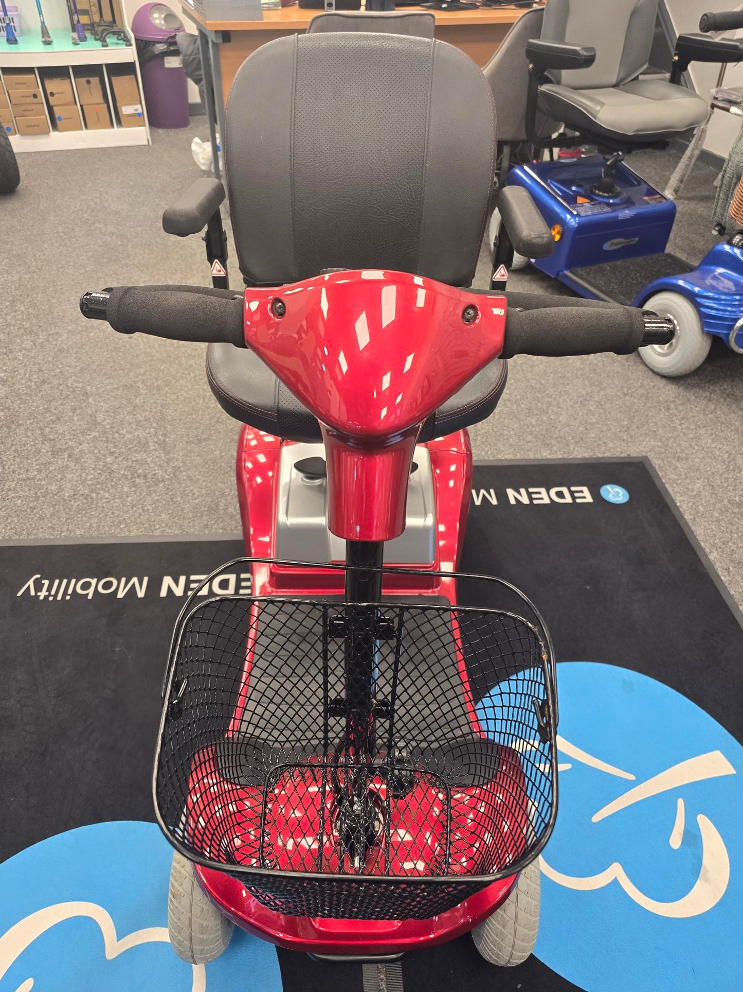 Red Kymco Mini Comfort mobility scooter with black seat, foam handles, and front basket on showroom floor mat