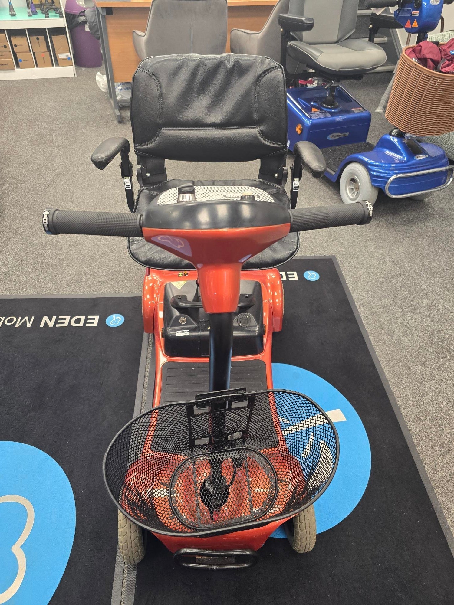Electric Mobility Ultralite 480 orange mobility scooter with black seat and front basket in indoor showroom setting