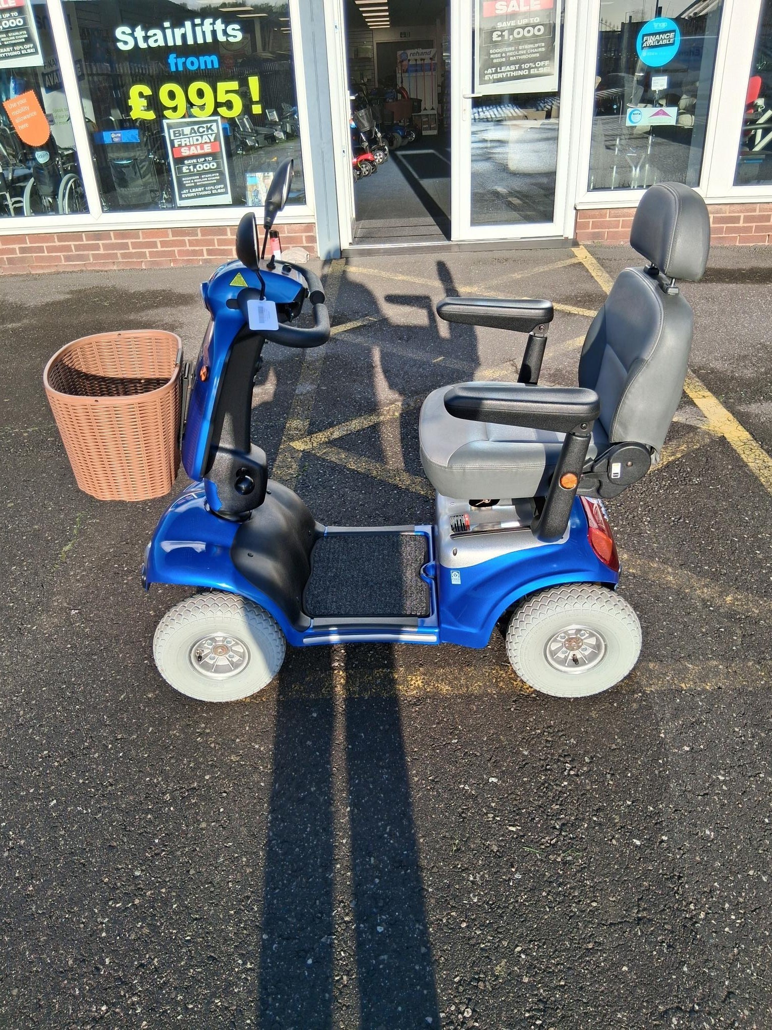 Blue Roma Shoprider Cadiz mobility scooter with black seat and armrests parked outside a store with a basket attached.
