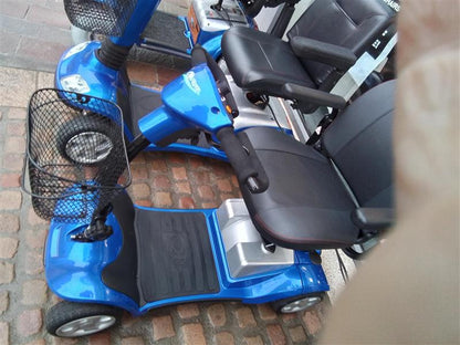 Blue Kymco Mini Comfort mobility scooter with black seat, armrests, front basket, and four wheels on cobblestone ground