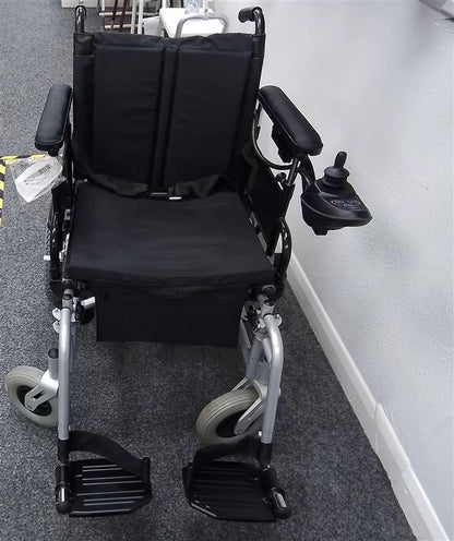 Black and grey powered wheelchair with cushioned seat, armrests, joystick control, and footrests on a gray carpeted floor