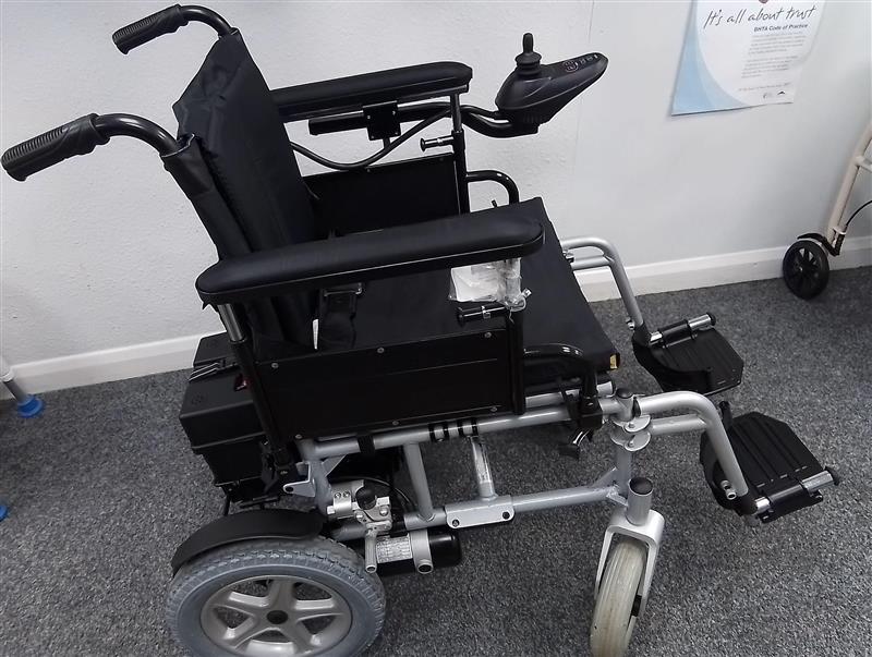 Black and grey Drive Cirrus powerchair with joystick control on gray carpeted floor indoors next to a wall poster