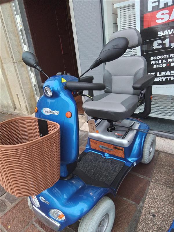 Blue Kymco Mini Comfort mobility scooter with grey seat, armrests, rearview mirrors, and a brown front basket outside a store