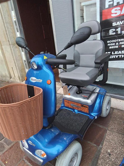 Blue Kymco Mini Comfort mobility scooter with grey seat, armrests, rearview mirrors, and a brown front basket outside a store