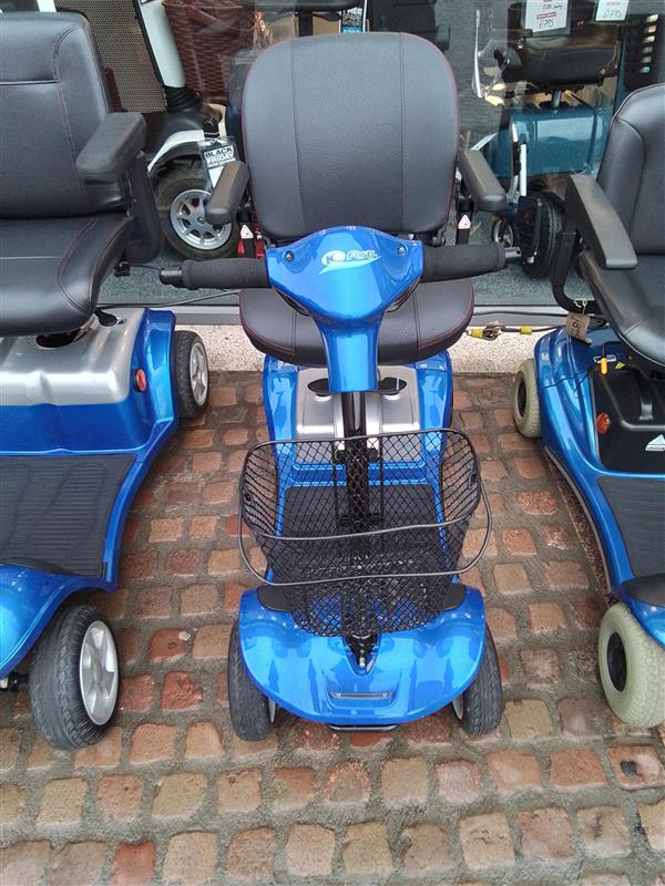 Blue Kymco Mini Comfort mobility scooter with black seat, armrests, and front basket parked on cobblestone pavement