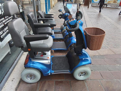 Blue Kymco Mini Comfort mobility scooter with black seat and basket parked on a tiled pedestrian street outside a store