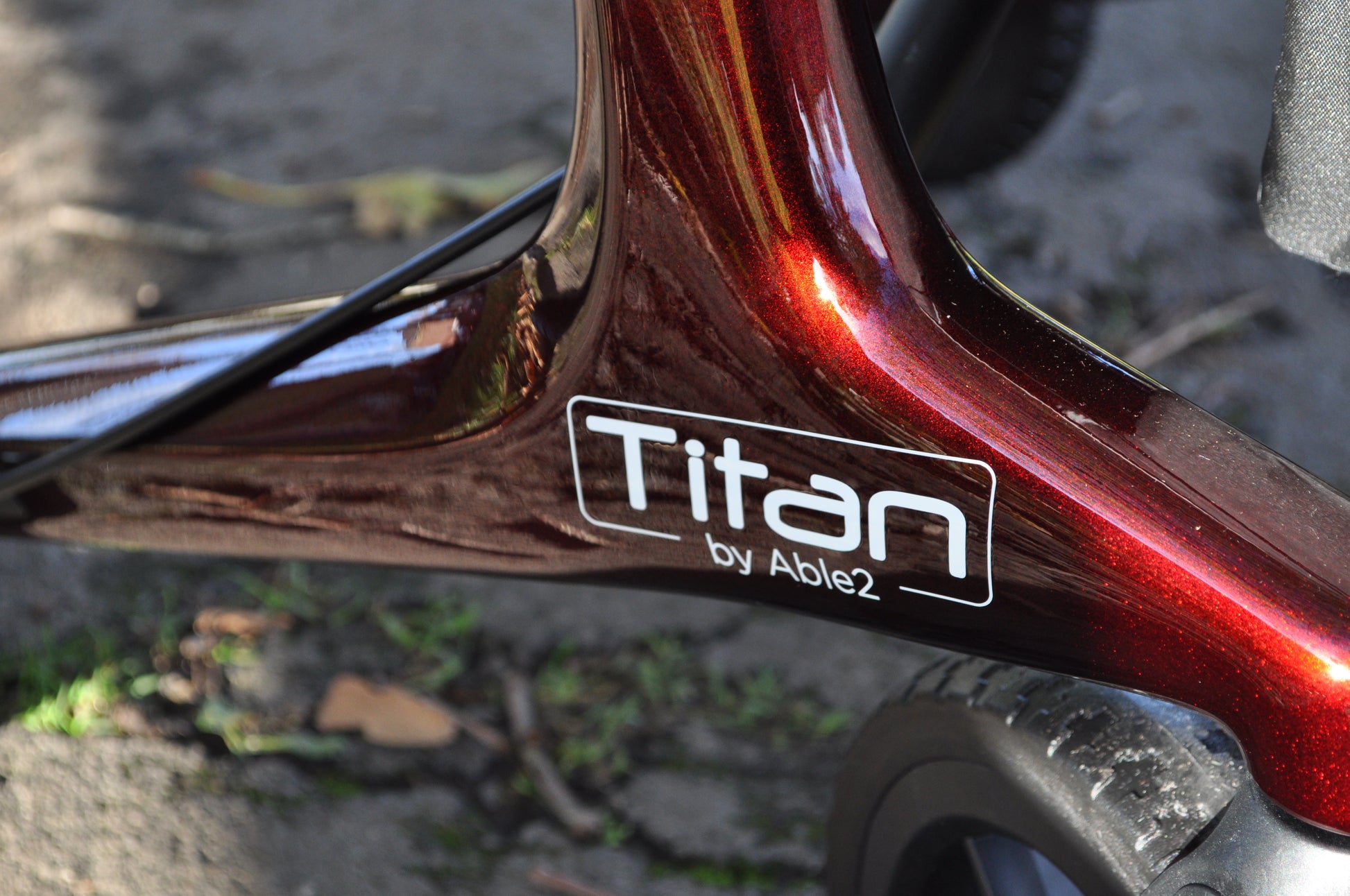 Close-up of deep red Able2 Titan Carbon Rollator frame with logo near black wheel on outdoor pavement