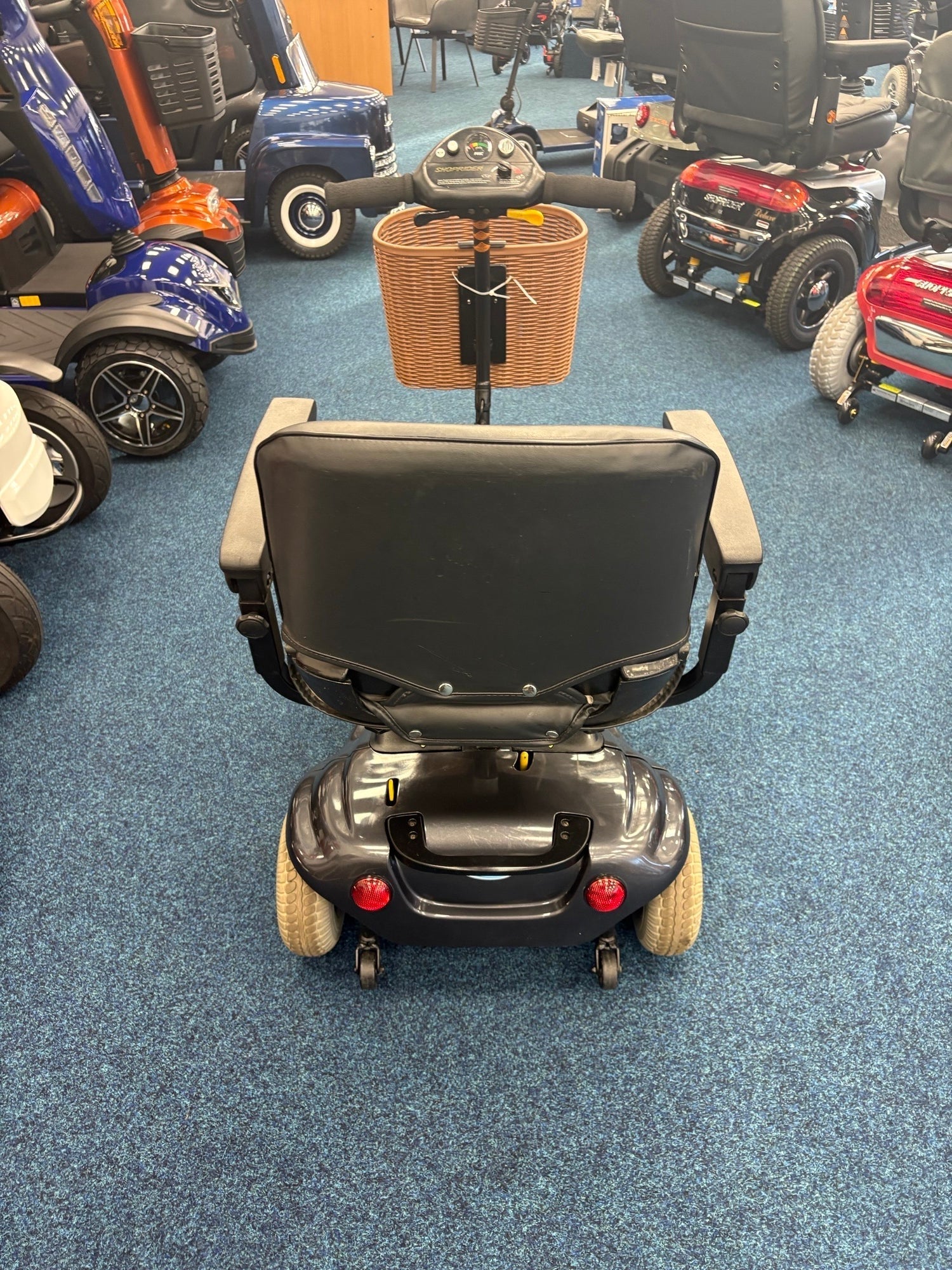 Rear view of refurbished Roma Paris Grey mobility scooter with basket, displayed indoors on blue carpet among other scooters