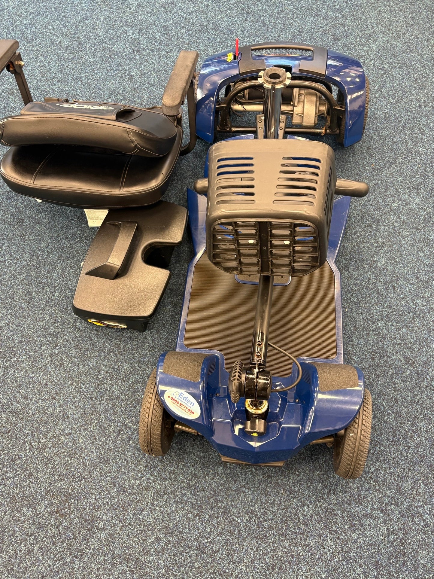 Disassembled Pride Apex Lite Blue mobility scooter with seat and battery pack on blue carpeted floor