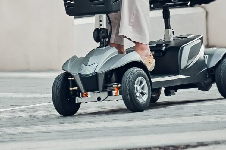 Close-up of a person riding The Eden Essential Aura 4mph boot mobility scooter on a paved surface outdoors