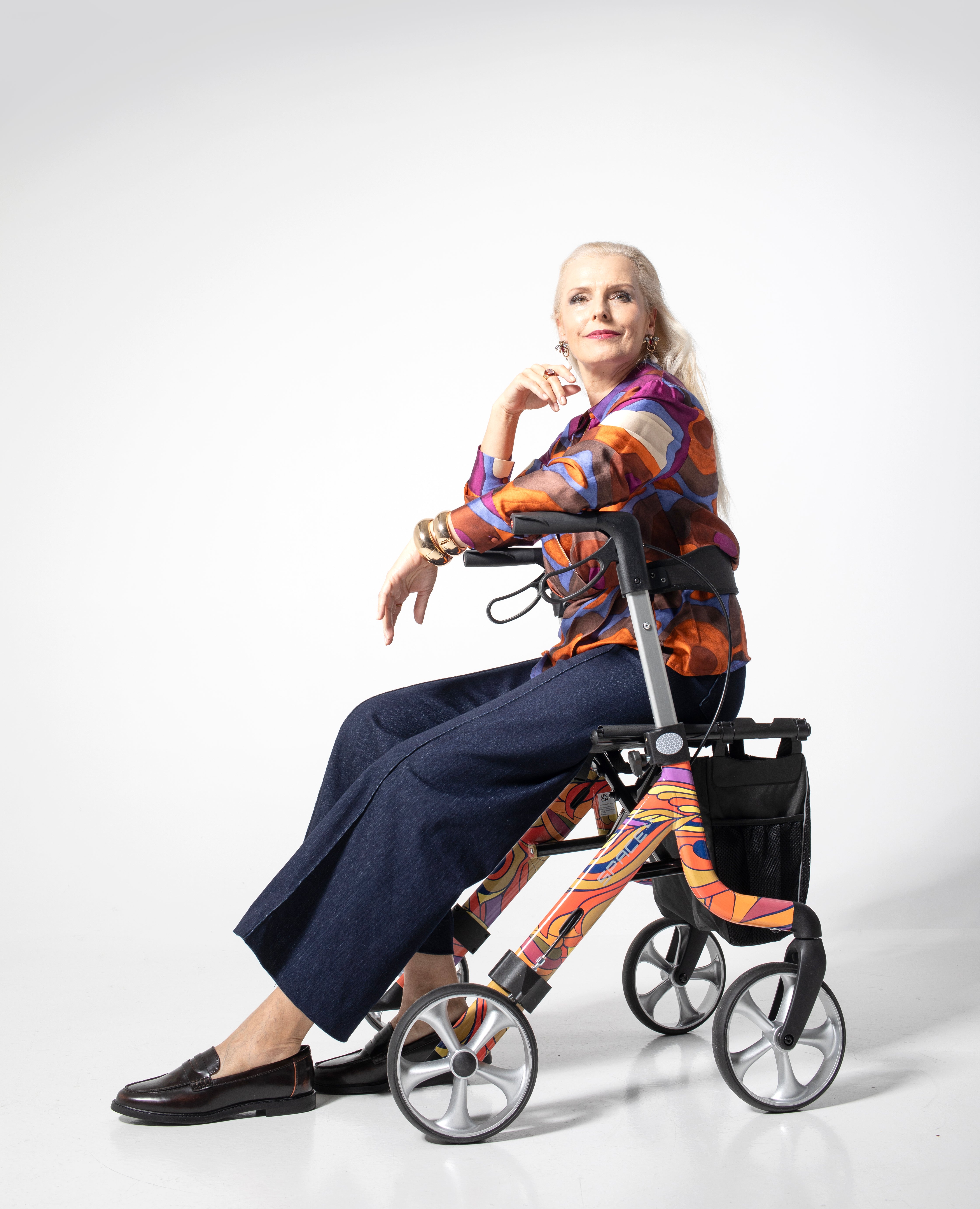Elderly woman sitting on a colorful Space LX Art Line Rollator walking aid with large wheels and storage pouch