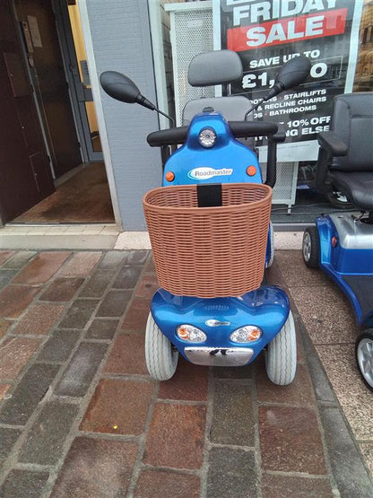 Blue Kymco Mini Comfort mobility scooter with brown front basket parked on stone pavement outside a store entrance.